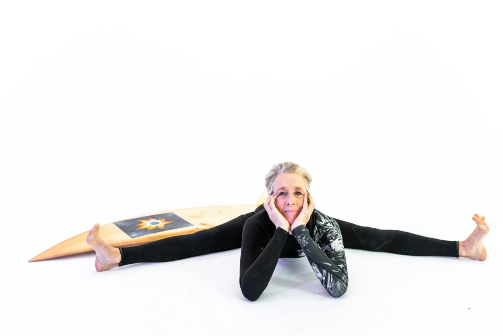 Cheerful senior woman in activewear stretching with a surfboard indoors, embracing vitality and wellness.
